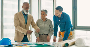 A team of professionals collaborating over a blueprint in a modern office, discussing estate and succession planning amidst color samples and architectural plans.