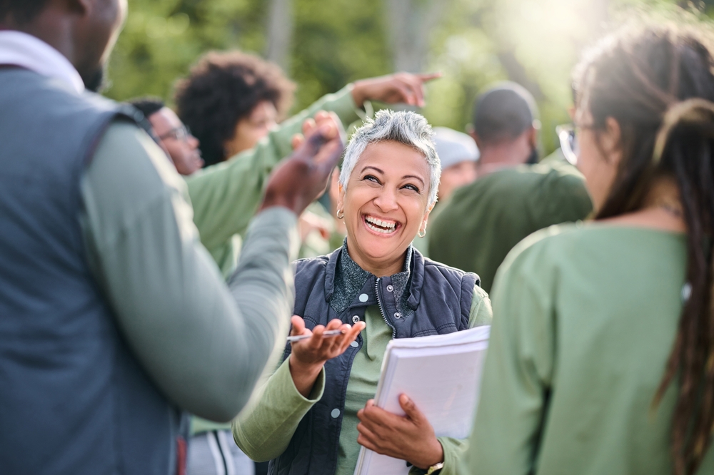A diverse group of volunteers engaging in a lively outdoor discussion, with a smiling woman holding documents at the center. The scene captures the spirit of teamwork and enthusiasm typical of philanthropic endeavors