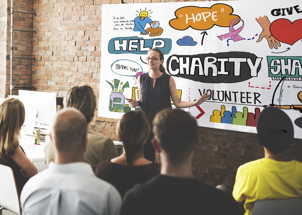 Corporate charity donations presentation in a modern office setting with a female speaker addressing a seated audience. The background features a colorful whiteboard illustration highlighting concepts like charity, help, hope, volunteer, and giving, emphasizing teamwork and philanthropic efforts.