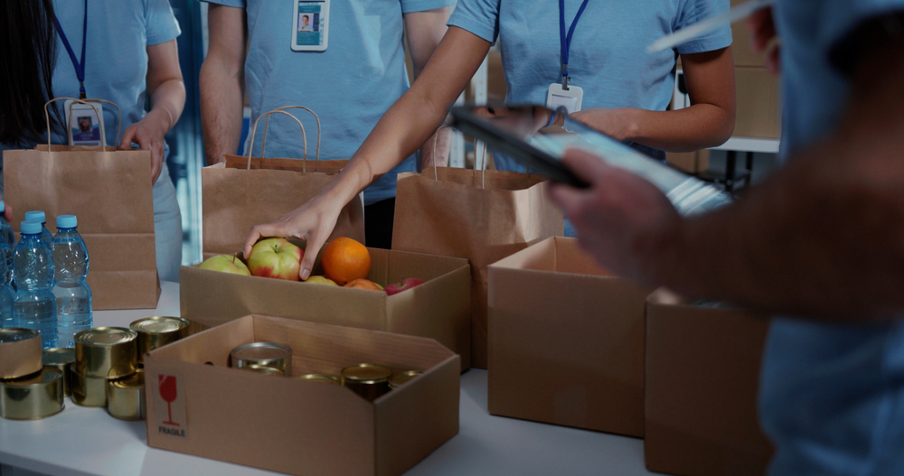 Volunteers packing food items and canned goods into boxes and bags at a donation center, representing community support and highlighting charitable giving demographics.
