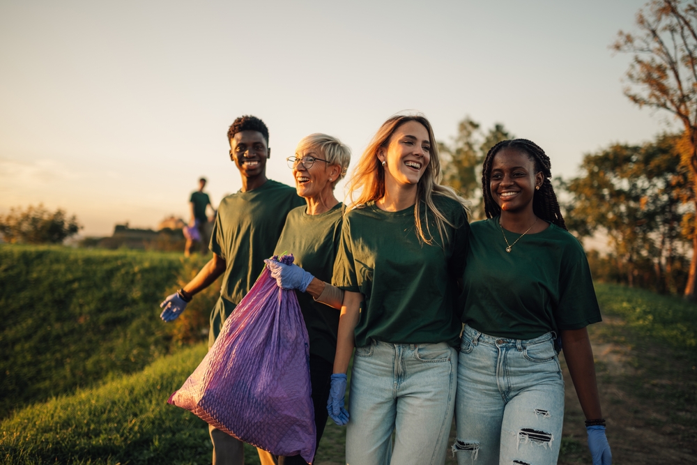 A diverse group of volunteers smiling while cleaning up outdoors, carrying a purple trash bag and wearing gloves during a community volunteering activity.