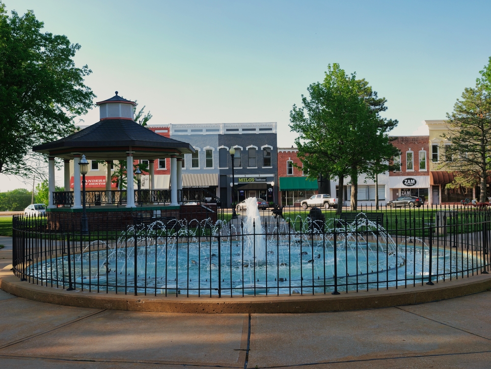 Small-town plaza with a fountain, gazebo, trees, and storefronts, representing community spaces and ways to give locally.