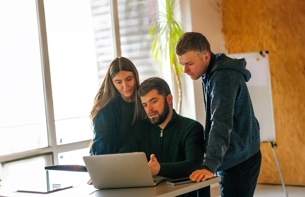 Three coworkers look at a laptop together in an office with a whiteboard behind them
