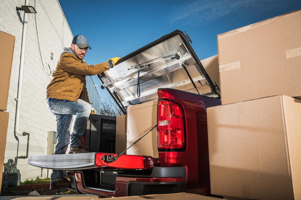 Man in a work jacket loading cardboard boxes into the bed of a red pickup truck