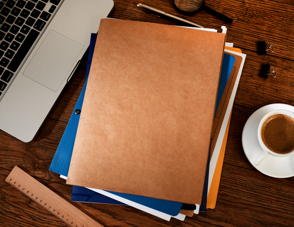 Manila and coloured document folders stacked on a wooden desk beside a laptop