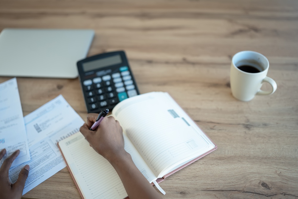 Hands writing in a planner beside a calculator, statements and coffee on a wooden table