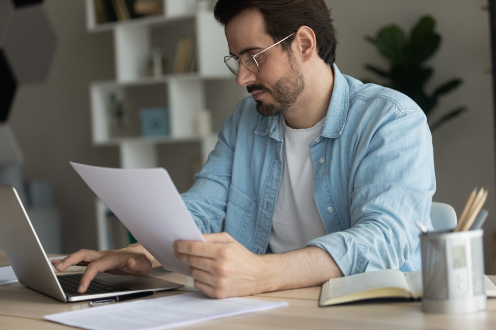 Man reading a printed document while typing on a laptop at his desk