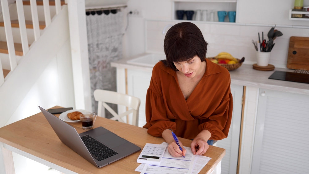Woman filling in a form at her kitchen table next to an open laptop