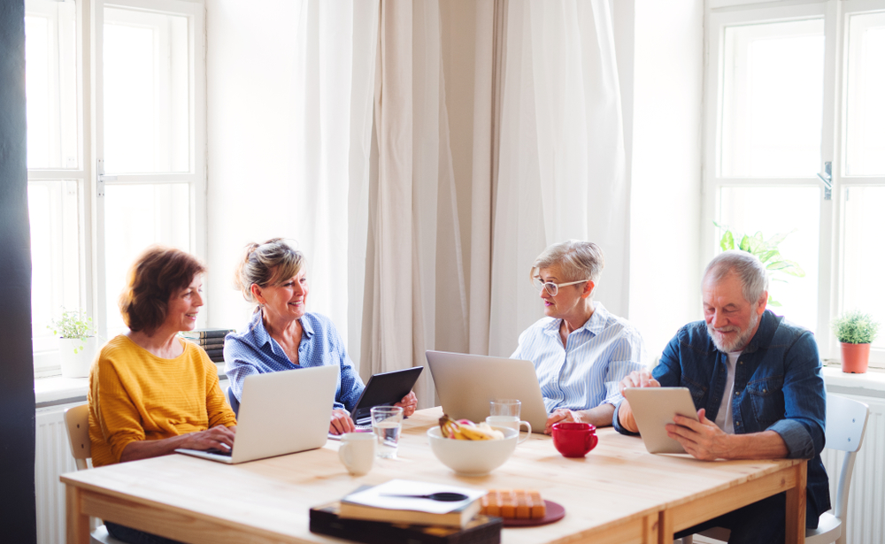 Four older adults working with laptops around a table in a bright community room