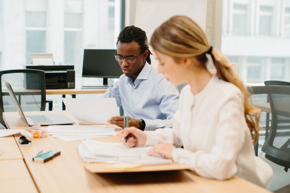 Two colleagues read through paperwork together at an office desk with a laptop
