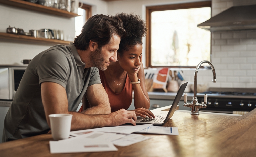 Two people review printed financial papers and a laptop at a sunlit kitchen counter