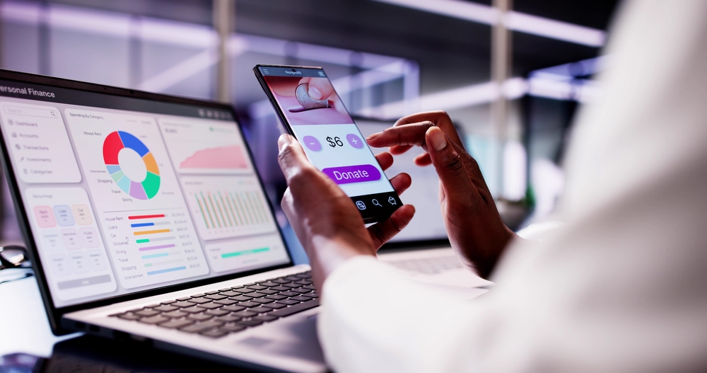 A person making an online donation by smartphone beside a laptop displaying financial charts, illustrating digital fundraising and donation management.