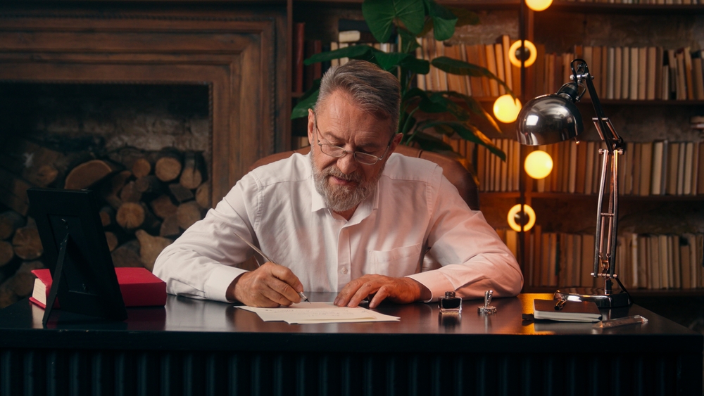 Older man writing a letter at a desk in a home office, illustrating in lieu of flowers wording for memorial notices and donation requests.