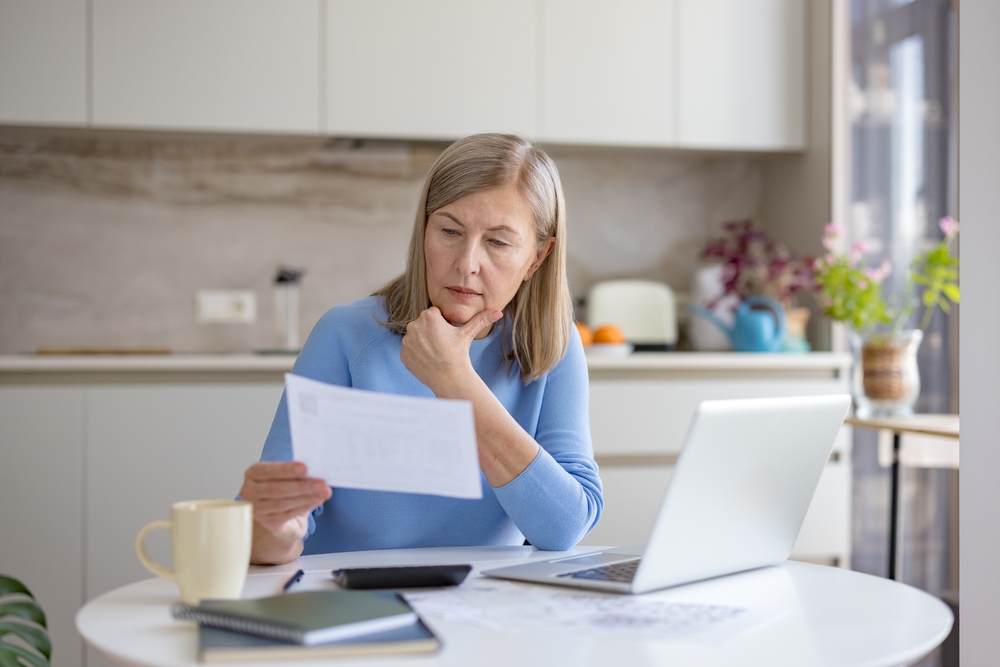 A woman in her seventies reviews paperwork at a kitchen table with a laptop open beside her