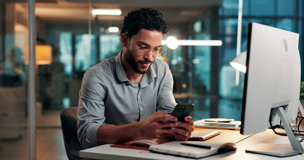 Professional using a smartphone at an office desk beside a computer and notebook, representing a grant proposal template workflow.