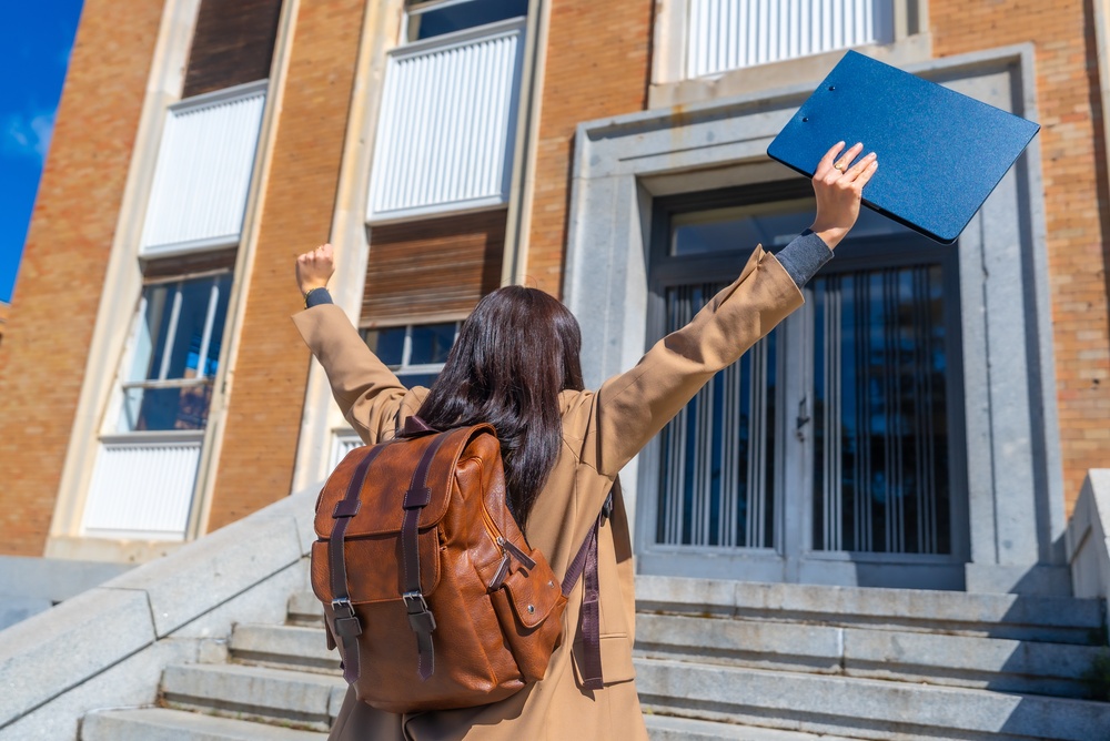 Small scholarships for high school seniors, showing a student with a backpack celebrating outside a school building while holding a folder overhead.