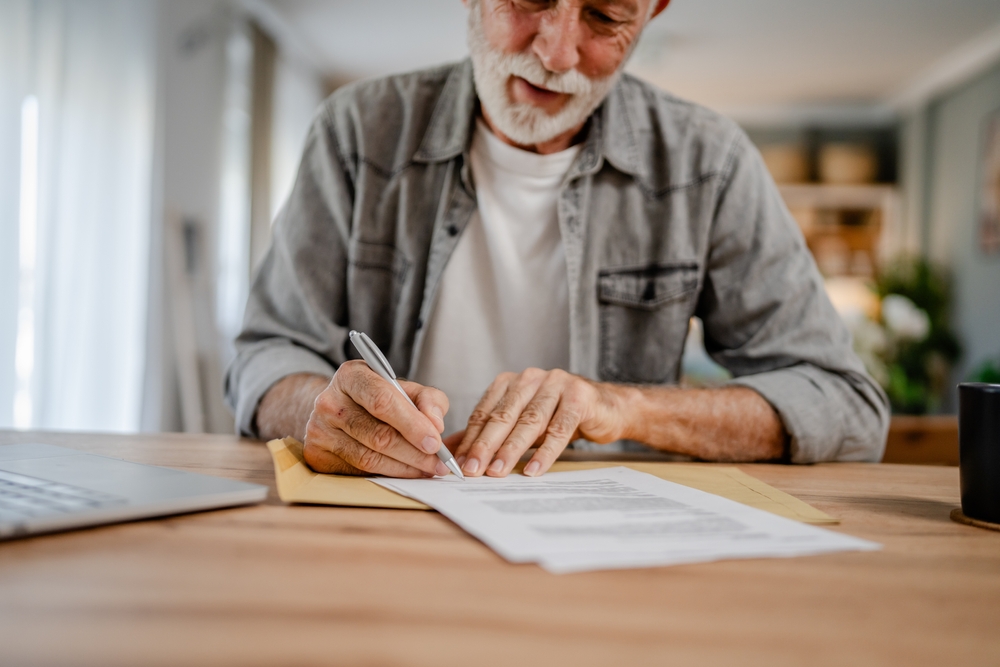 Older man signing a printed form at a wooden desk beside a laptop
