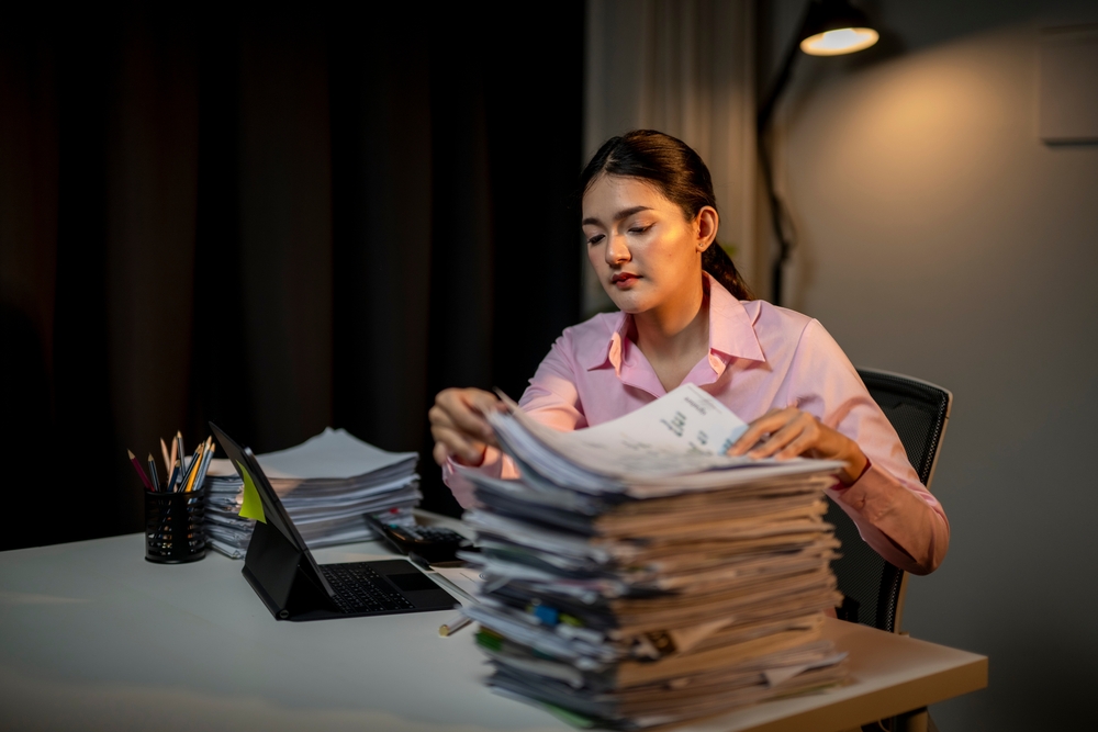 Woman sorting through a stack of papers alone at a desk under a lamp