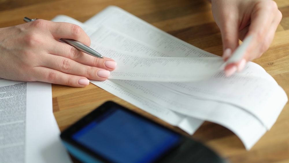 Hands reviewing printed financial documents with a pen on a wooden desk beside a tablet