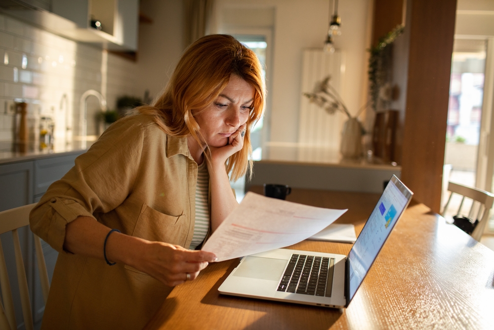 Woman at a kitchen table reading a printed financial statement next to an open laptop