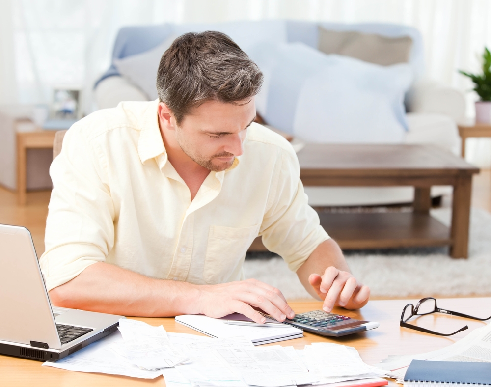 A man works through printed statements with a calculator and laptop at a home table