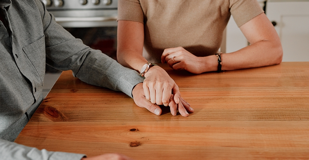 One person rests a hand over another's across a wooden kitchen table