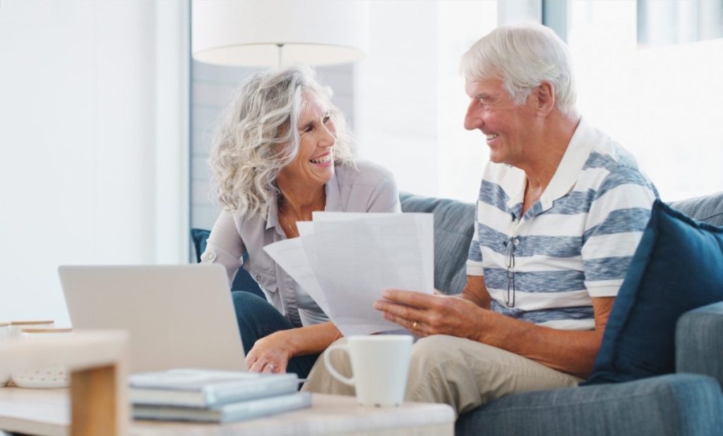 A smiling older couple reviewing paperwork beside a laptop at home, learning how to read a Form 990 before they give.