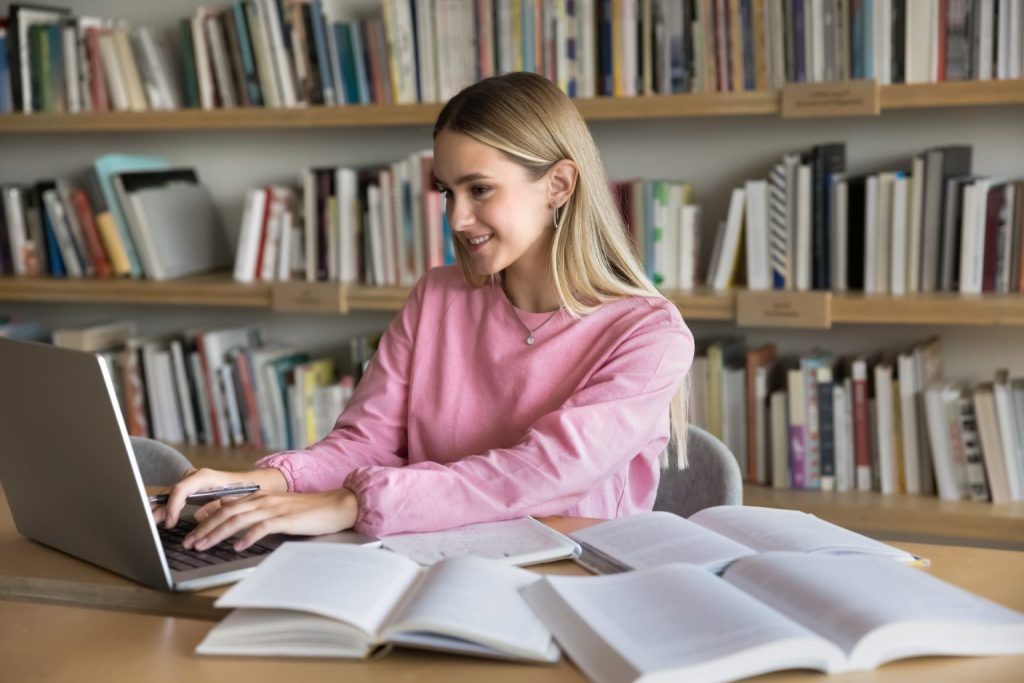 Smiling student typing at a laptop in a library, ready to write a scholarship essay.