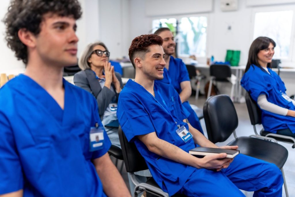 Nursing students in blue scrubs listen to an instructor in a classroom, the training many nursing scholarships in Kansas help pay for.