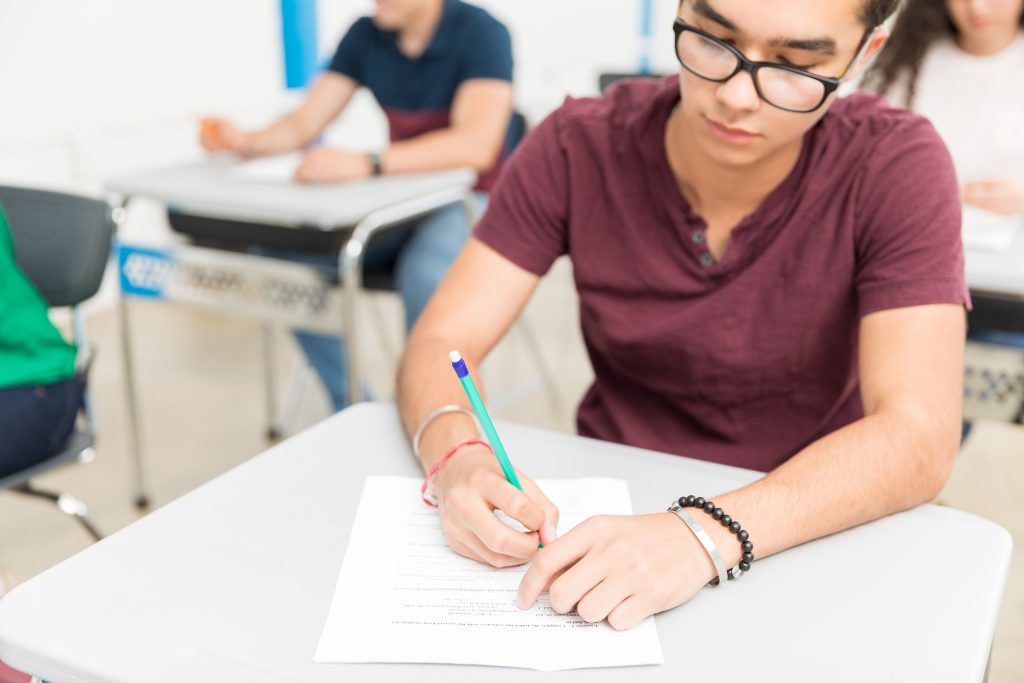 Student writing with a pencil at a classroom desk, starting to write a scholarship essay from the real prompt.